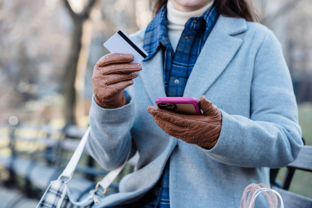 Crop unrecognizable female in casual clothes and gloves browsing mobile phone and entering credit card details while sitting on bench in park in daylight